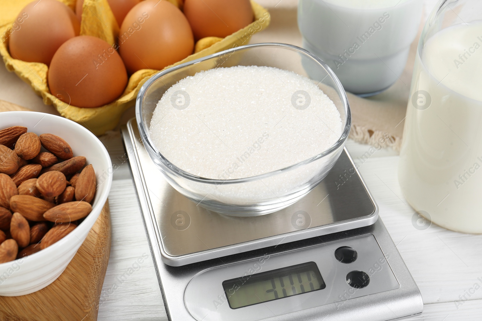 Photo of Kitchen scale with bowl of sugar among milk, almond and eggs on white wooden table, closeup