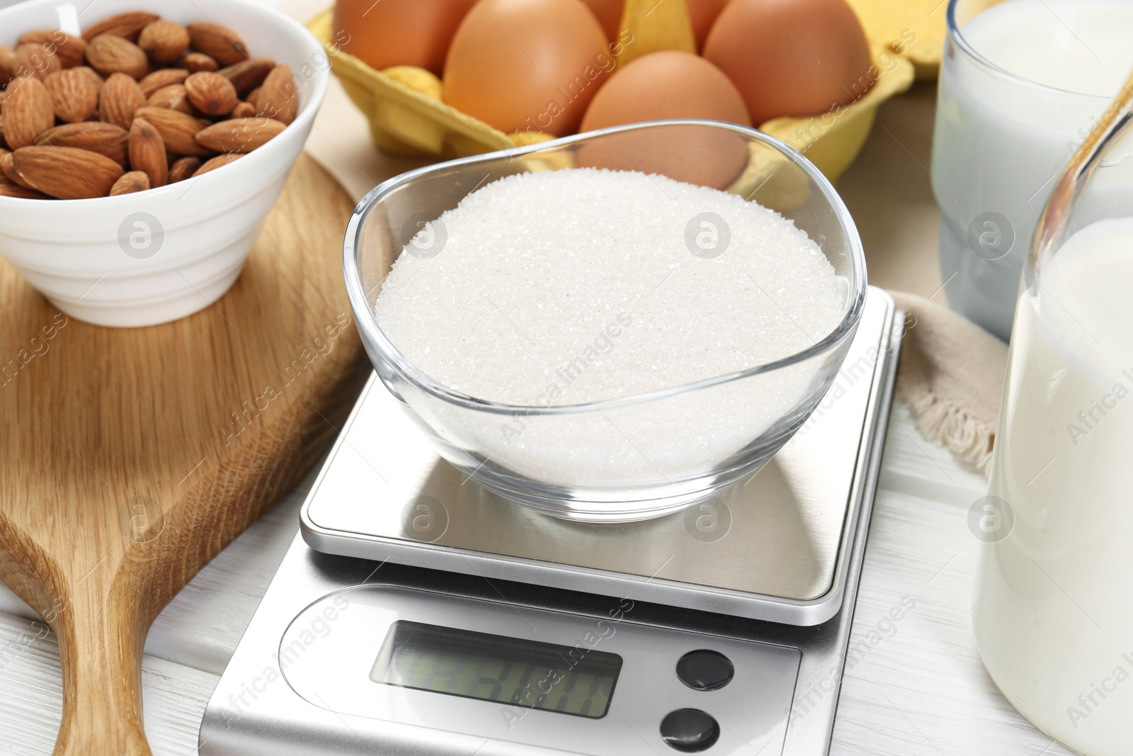 Photo of Kitchen scale with bowl of sugar among milk, almond and eggs on white wooden table, closeup