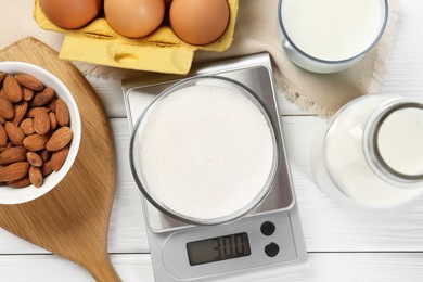 Kitchen scale with bowl of sugar among milk, almond and eggs on white wooden table, flat lay