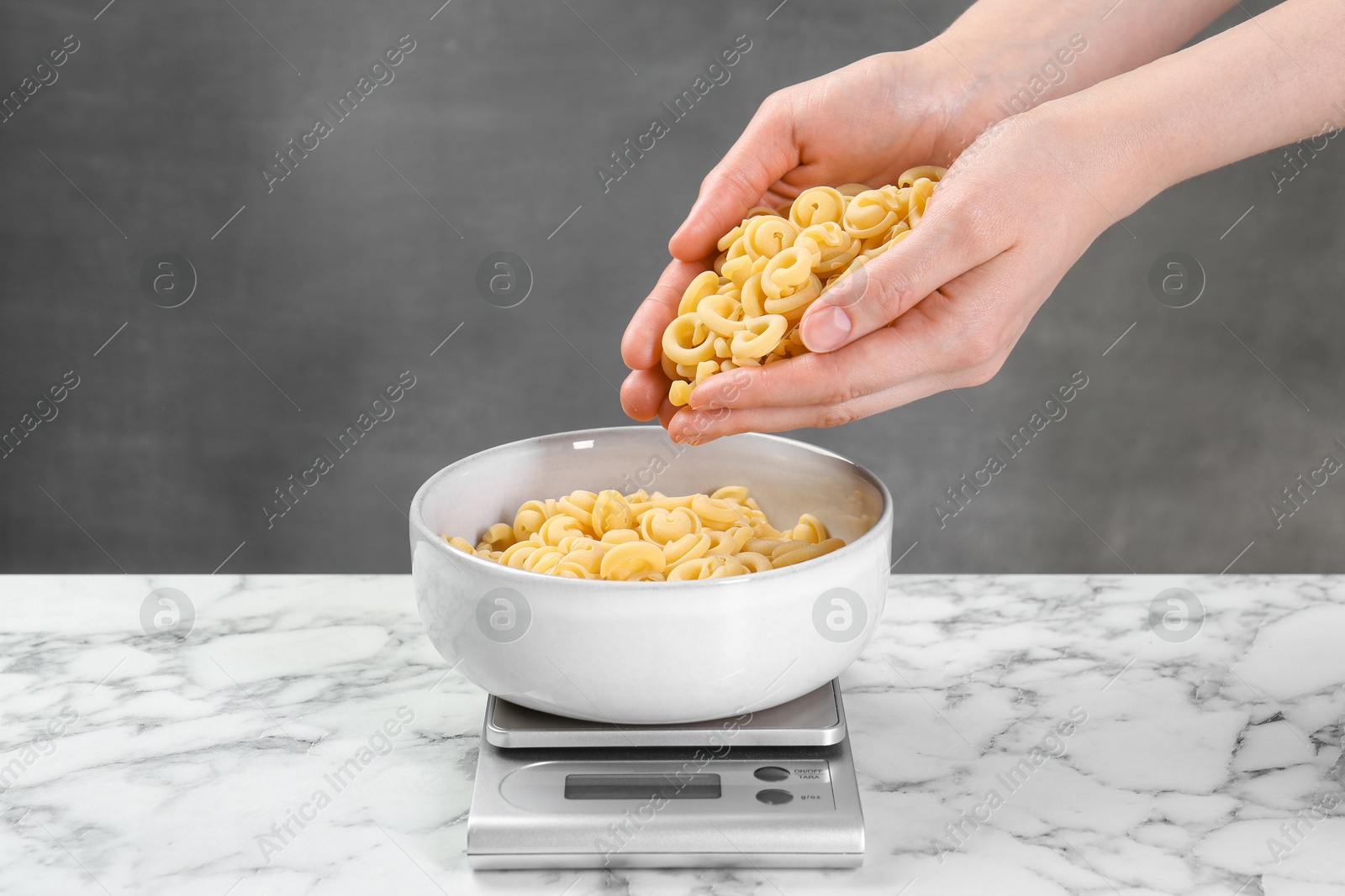 Photo of Woman adding pasta into bowl on kitchen scale on white marble table, closeup