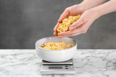 Woman adding pasta into bowl on kitchen scale on white marble table, closeup