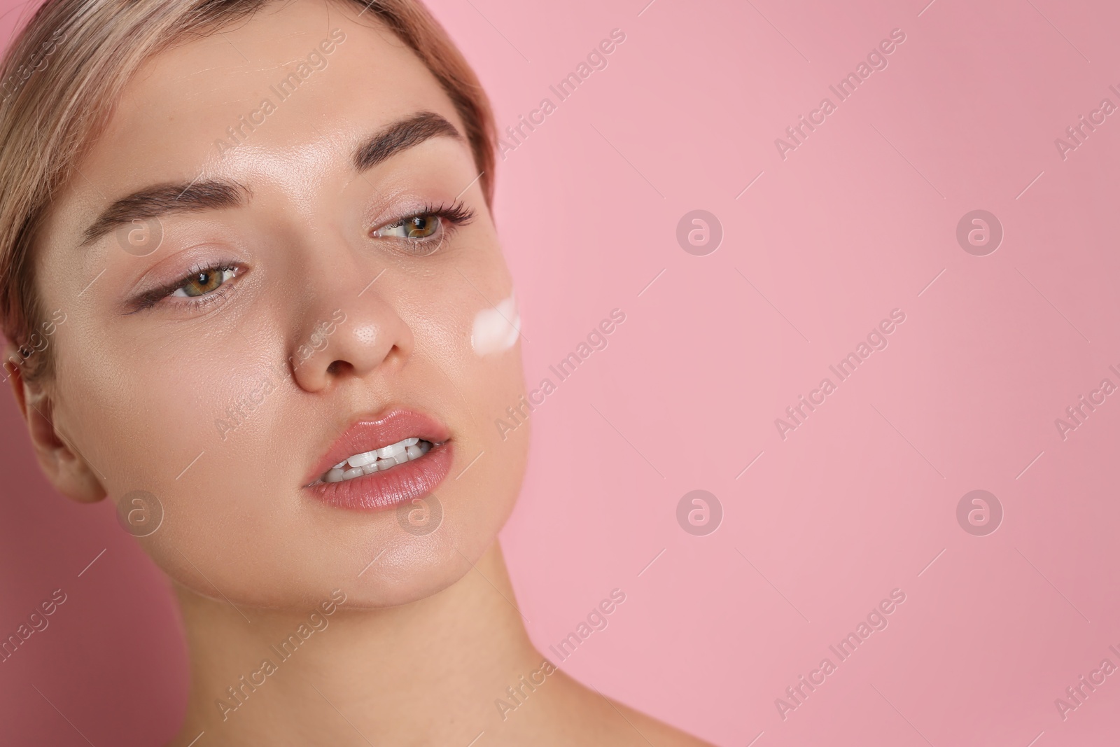 Photo of Beautiful woman with cream on her face against pink background, closeup. Space for text