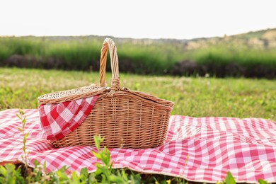 Photo of Picnic wicker basket with napkin and red checkered blanket on green grass outdoors, space for text