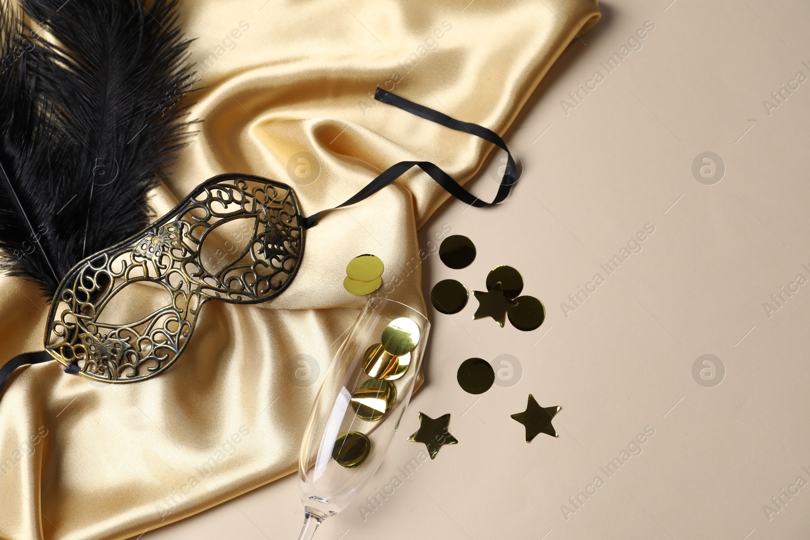 Photo of Beautiful carnival mask, champagne glass, confetti and silk fabric on beige background, flat lay. Space for text