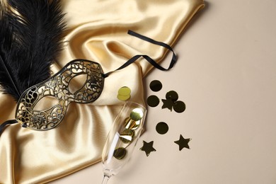 Beautiful carnival mask, champagne glass, confetti and silk fabric on beige background, flat lay. Space for text