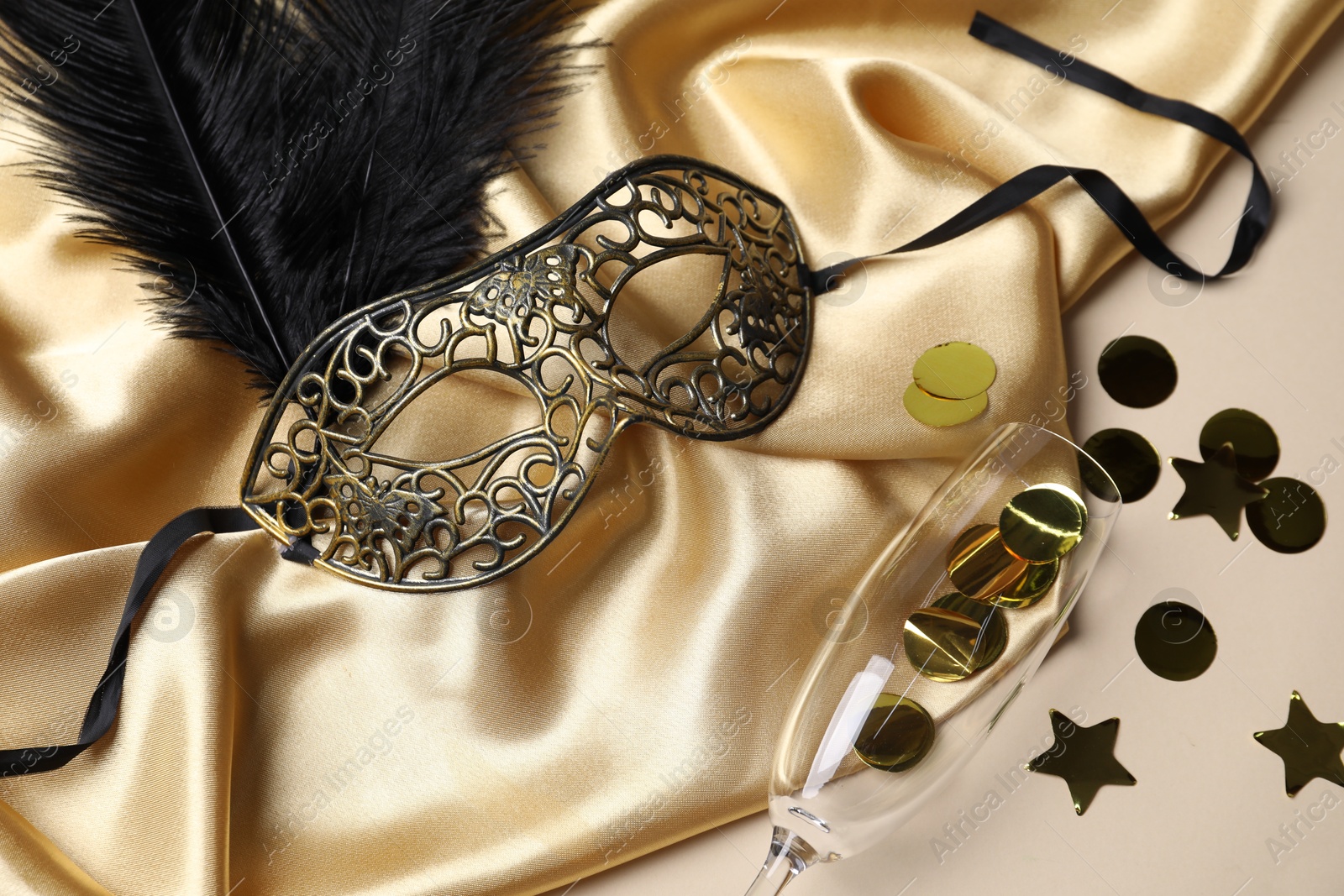 Photo of Beautiful carnival mask, champagne glass, confetti and silk fabric on beige background