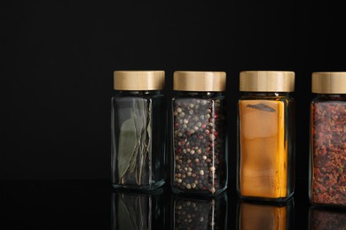 Different spices in glass jars on black mirror surface, space for text