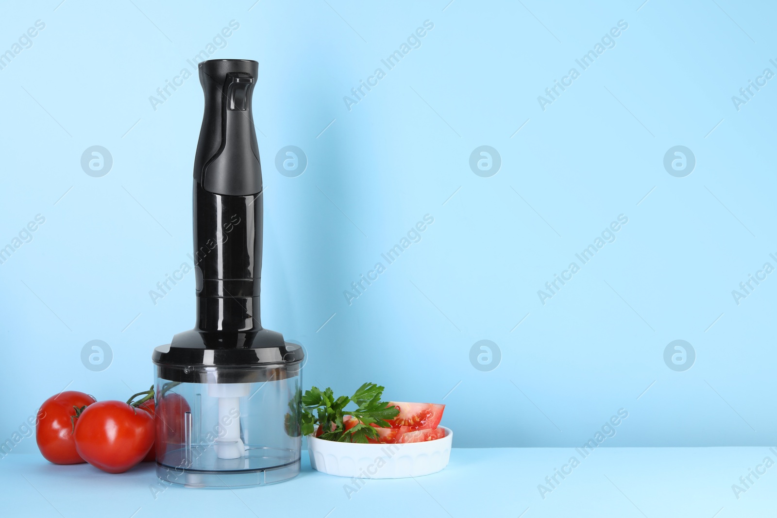 Photo of Hand blender kit, fresh tomatoes and parsley on light blue background, space for text