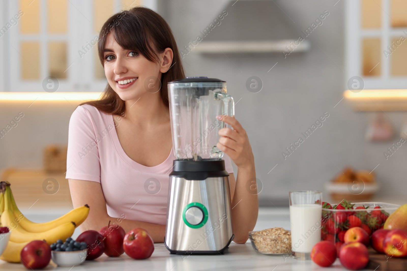 Photo of Young woman making delicious smoothie with blender at white marble table in kitchen