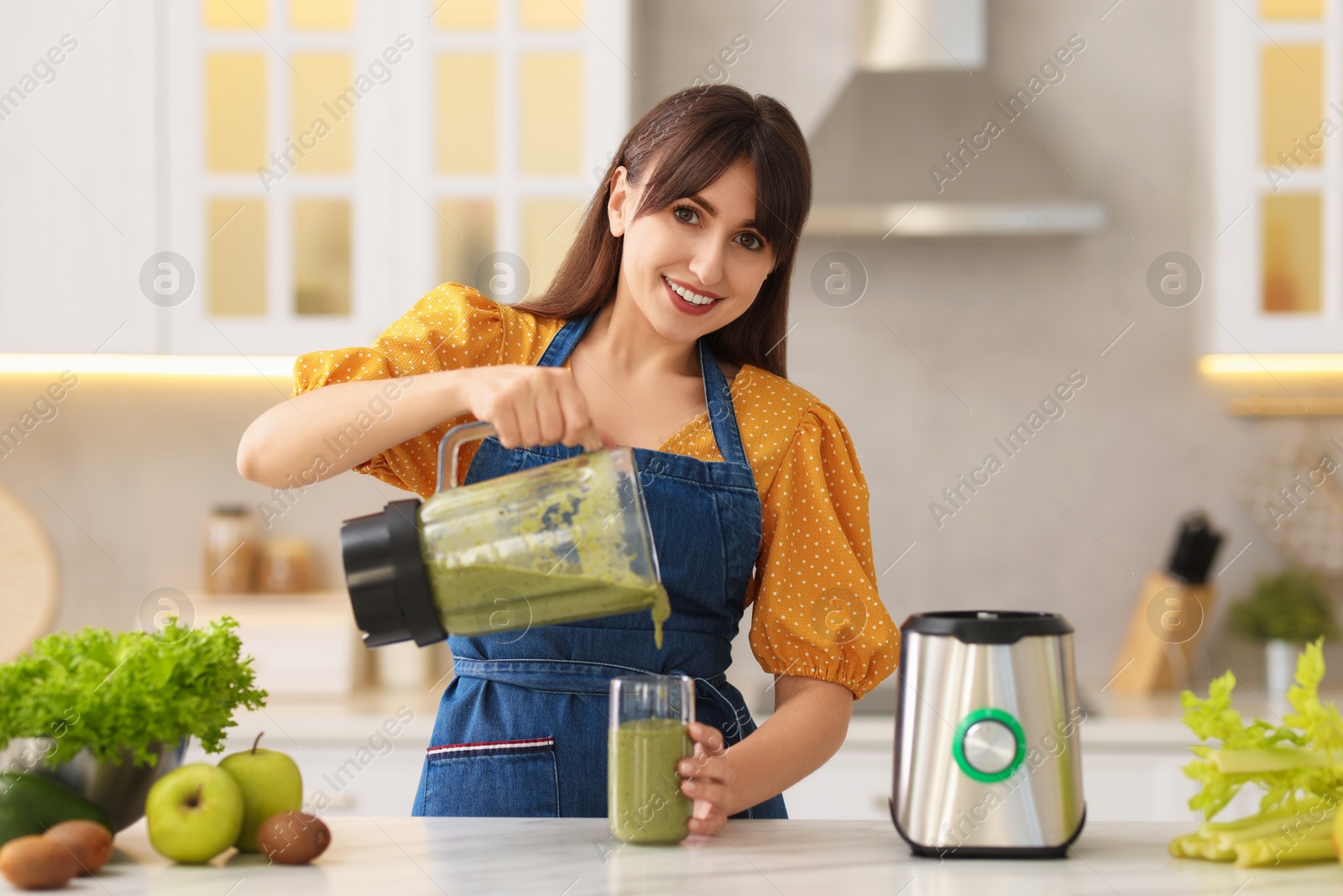 Photo of Young woman pouring fresh smoothie from blender cup into glass at white marble table in kitchen