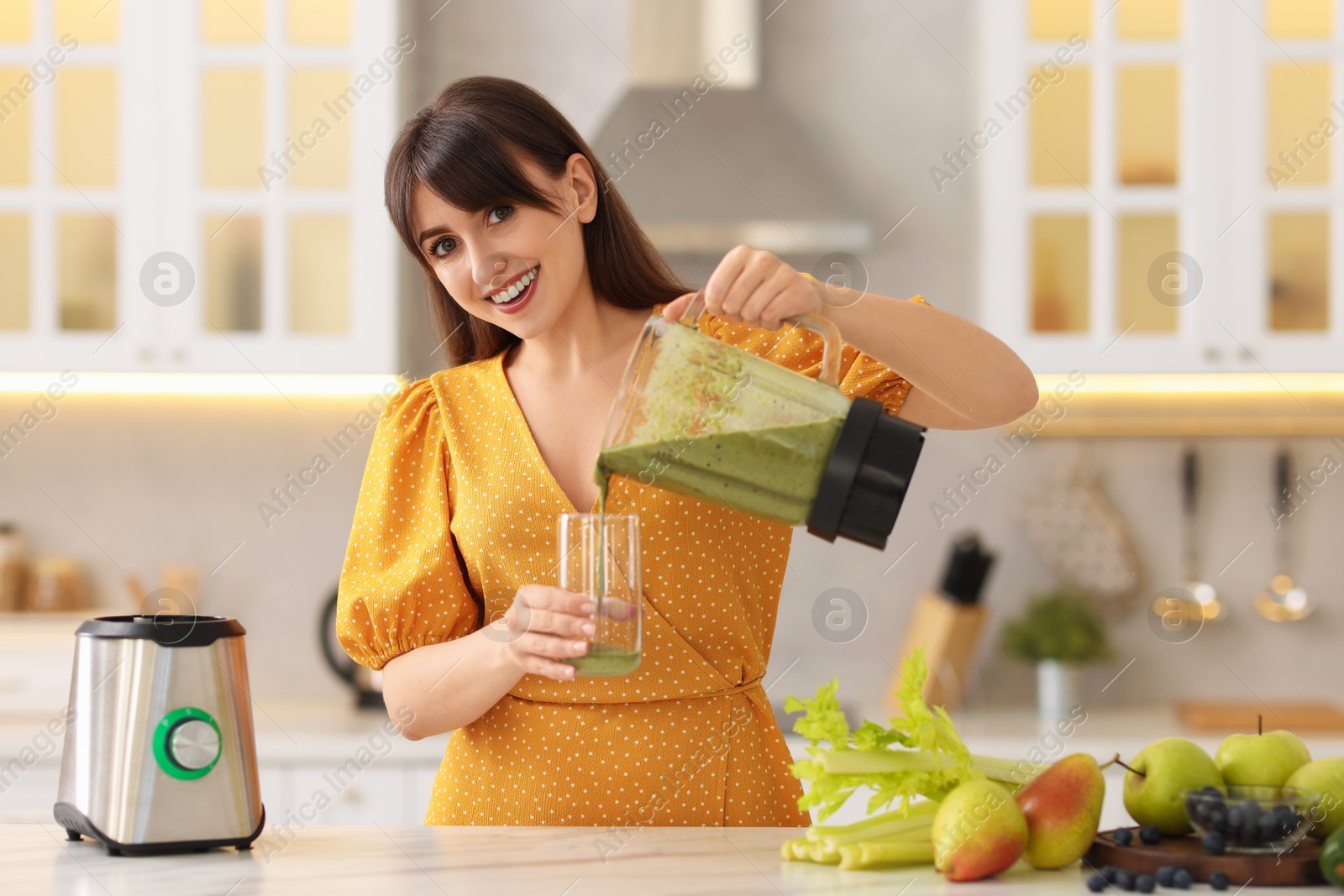 Photo of Young woman pouring fresh smoothie from blender cup into glass at white marble table in kitchen