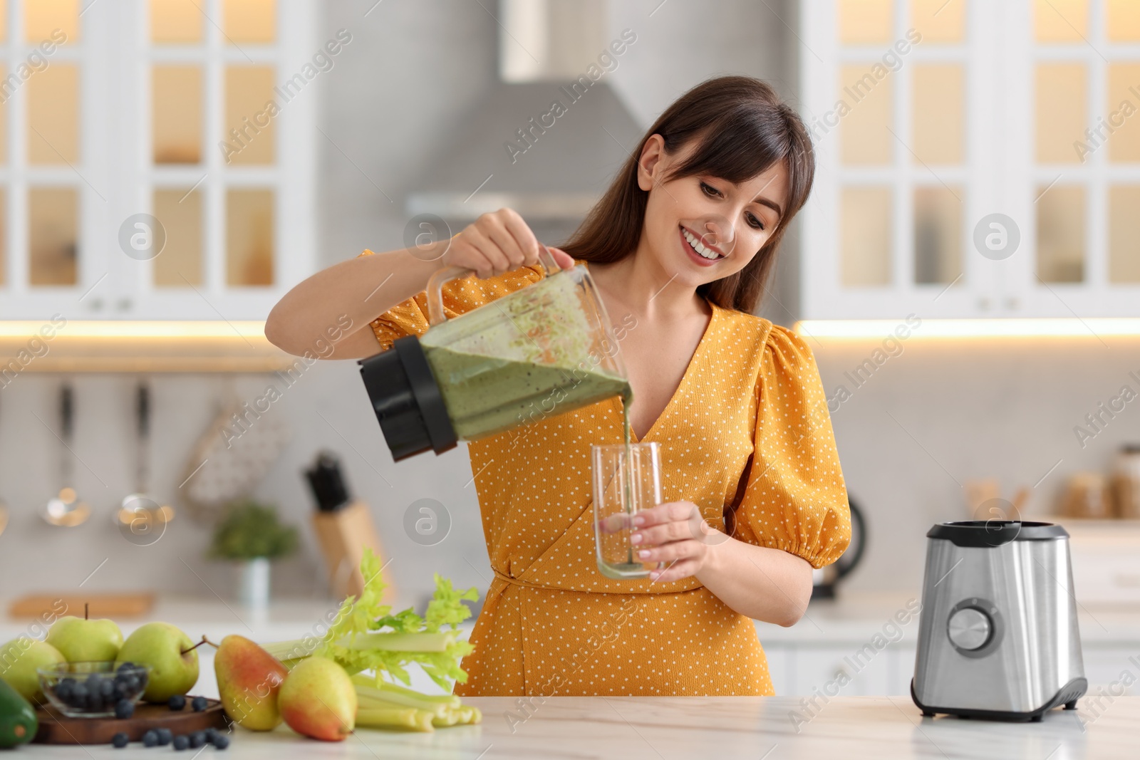 Photo of Young woman pouring fresh smoothie from blender cup into glass at white marble table in kitchen