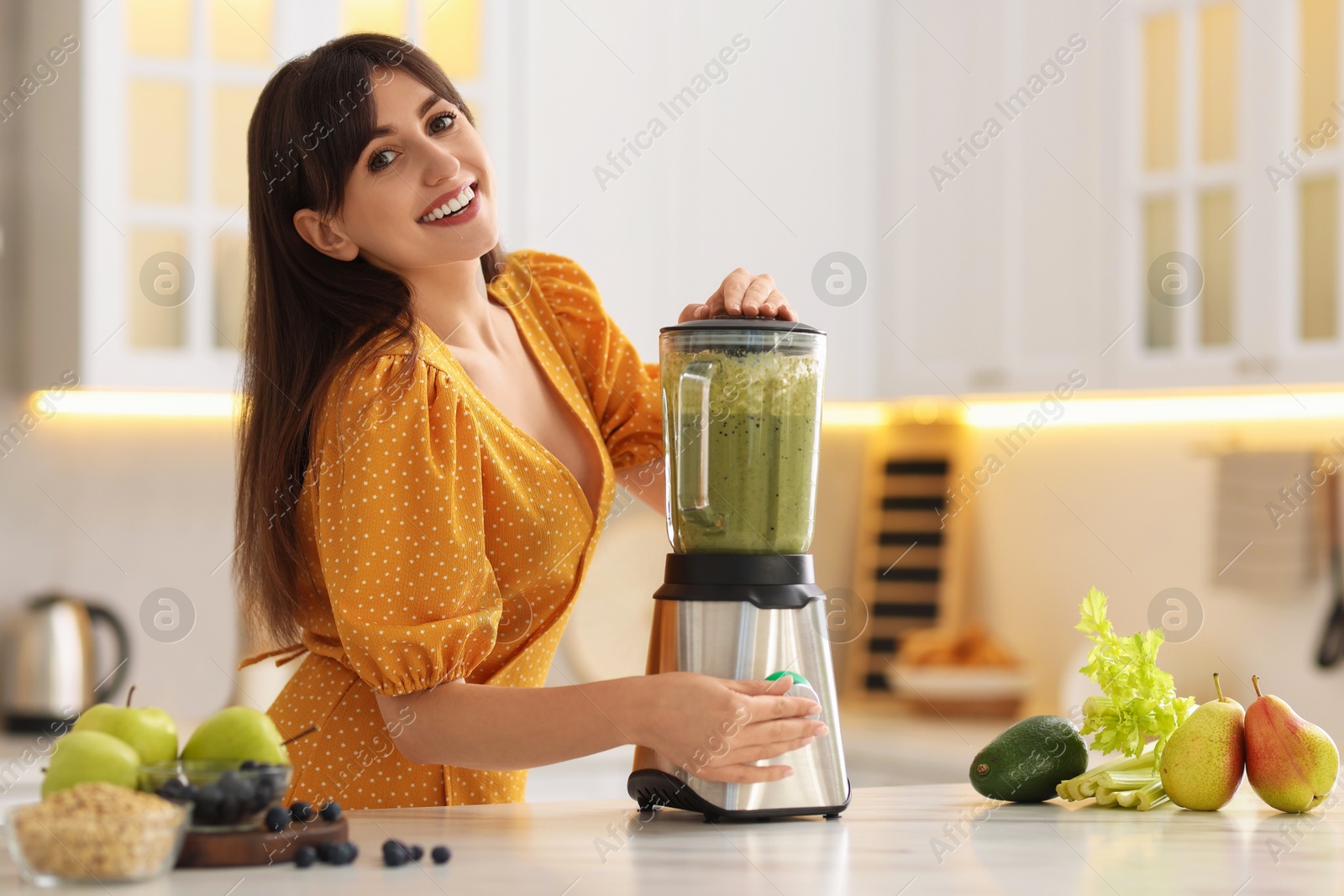 Photo of Young woman making delicious smoothie with blender at white marble table in kitchen