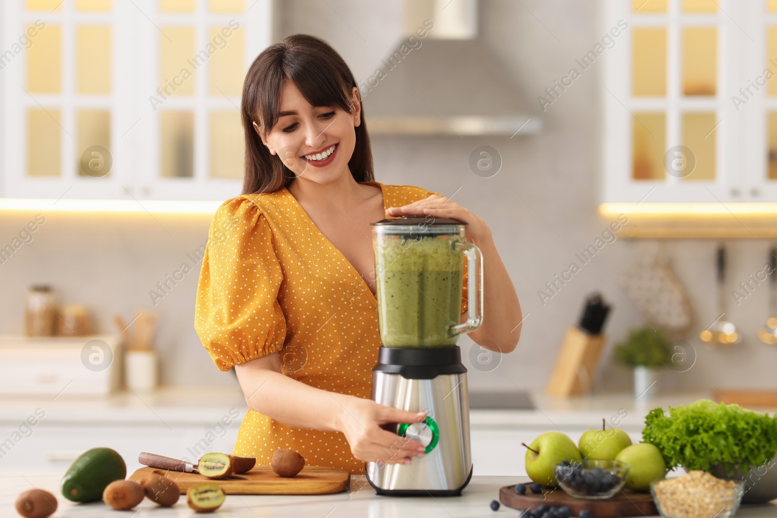 Photo of Young woman making delicious smoothie with blender at white marble table in kitchen