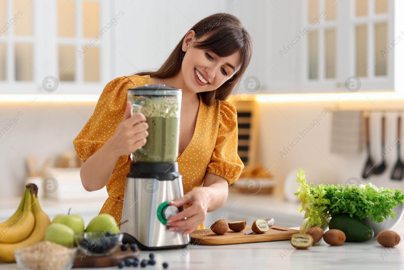 Photo of Young woman making delicious smoothie with blender at white marble table in kitchen