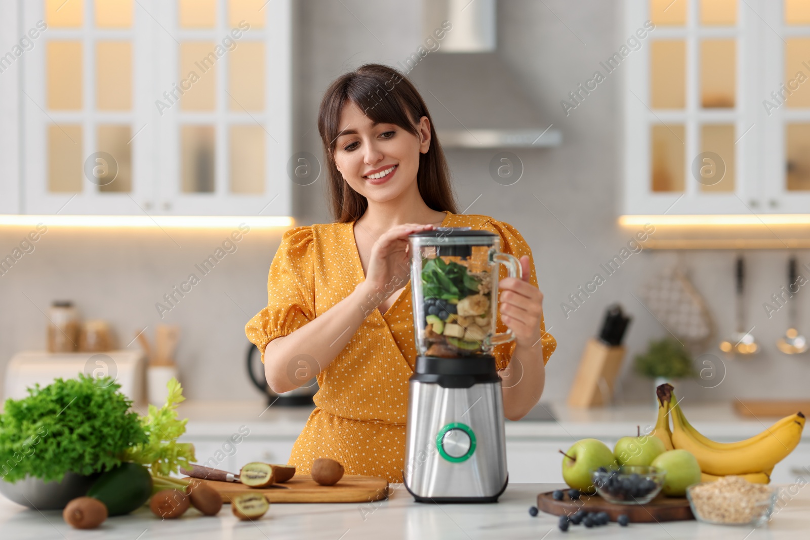 Photo of Young woman making delicious smoothie with blender at white marble table in kitchen