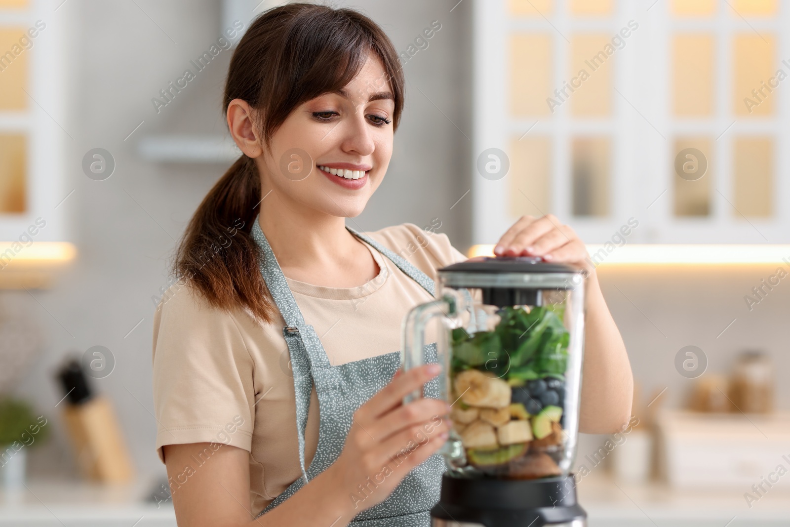 Photo of Young woman making delicious smoothie with blender in kitchen