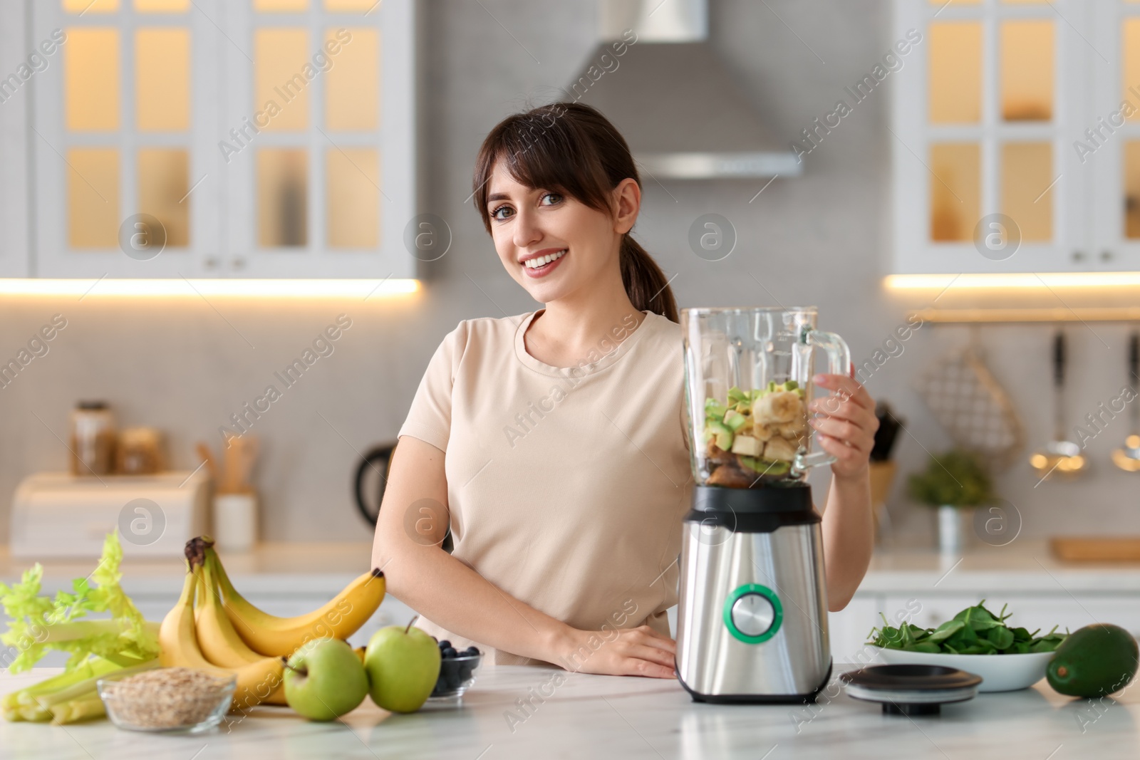 Photo of Young woman making delicious smoothie with blender at white marble table in kitchen