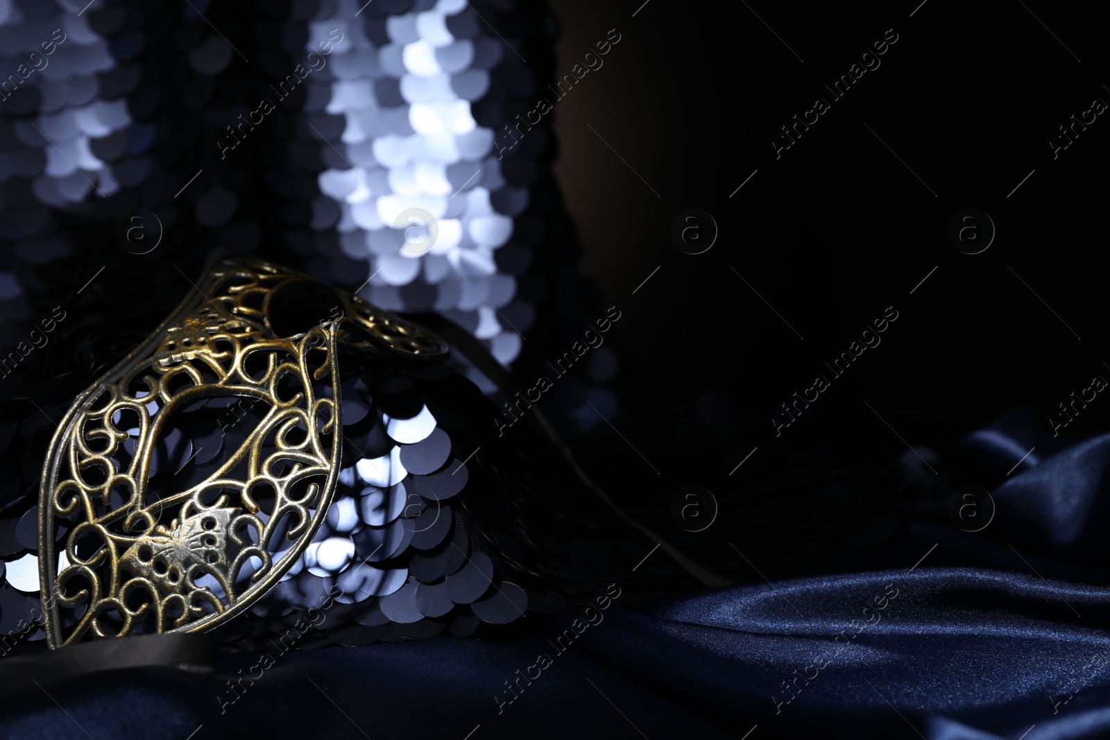 Photo of Beautiful carnival mask and fabric with golden sequins in darkness, closeup. Space for text