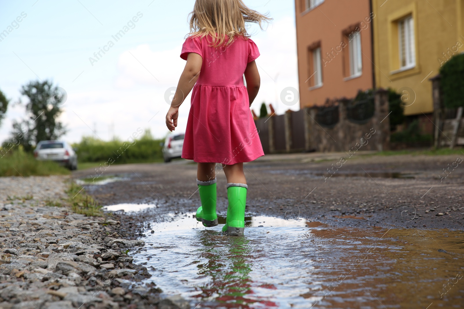 Photo of Little girl wearing green rubber boots walking in puddle outdoors, back view