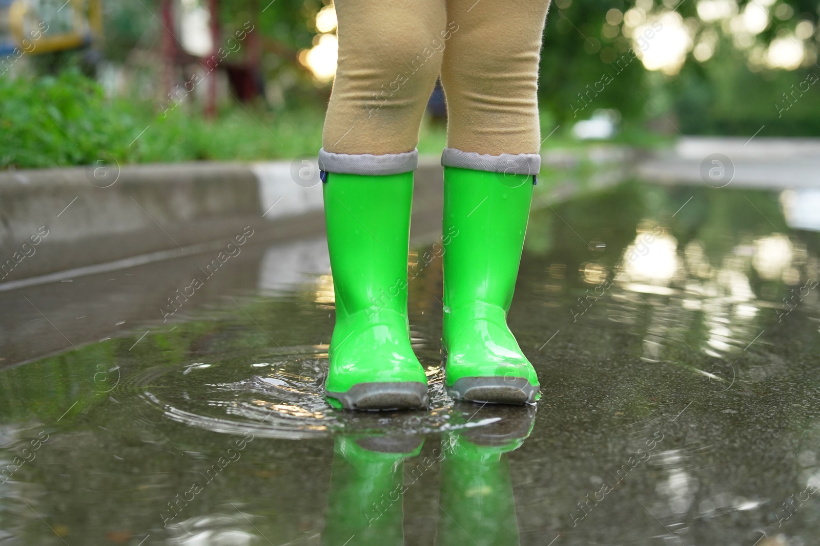 Photo of Little girl wearing green rubber boots standing in puddle outdoors, closeup