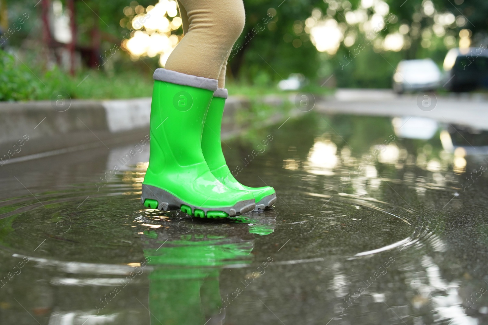 Photo of Little girl wearing green rubber boots standing in puddle outdoors, closeup