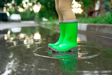 Photo of Little girl wearing green rubber boots standing in puddle outdoors, closeup