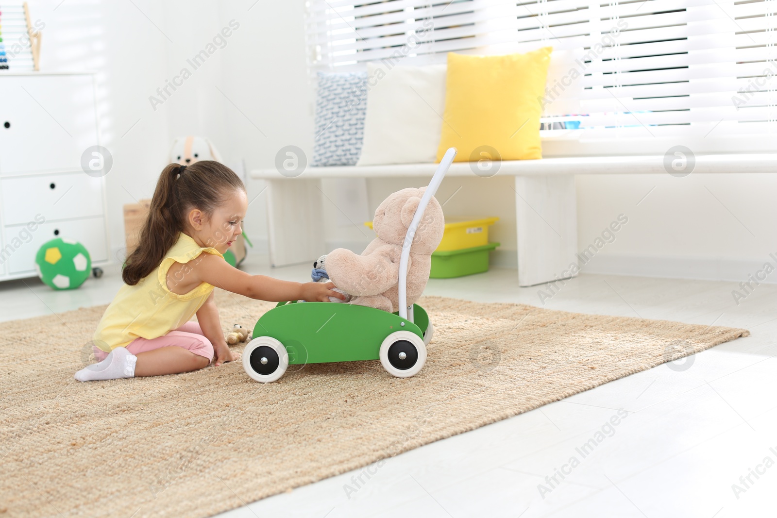 Photo of Cute little girl playing with toy walker and teddy bear at home