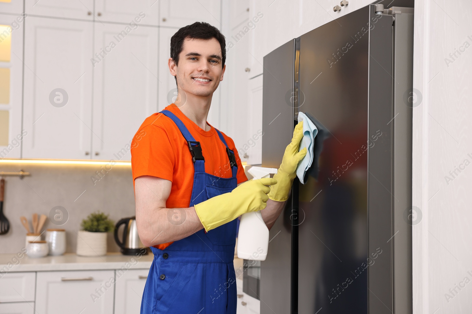 Photo of Professional janitor wearing uniform cleaning fridge in kitchen