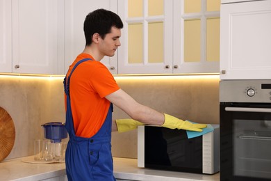 Photo of Professional janitor wearing uniform cleaning microwave oven in kitchen