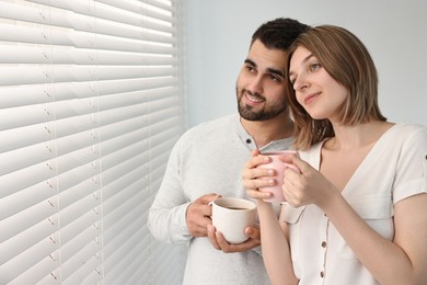 Photo of Couple with cups of drink near window blinds at home, space for text