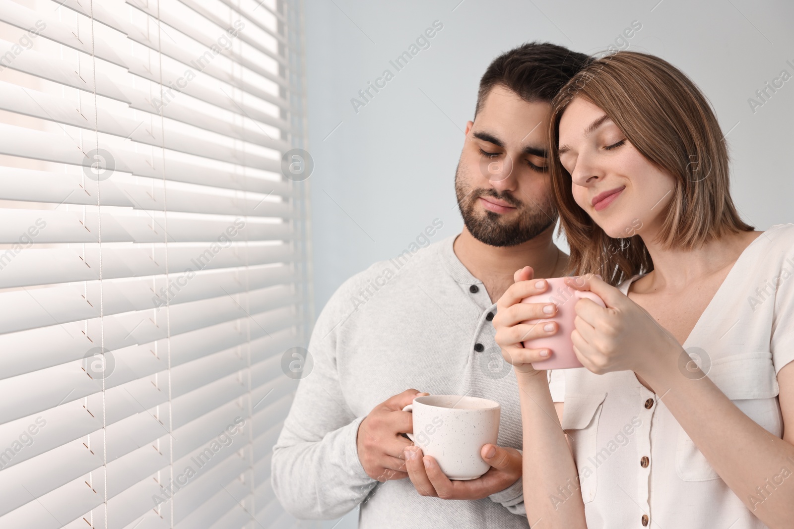 Photo of Couple with cups of drink near window blinds at home, space for text