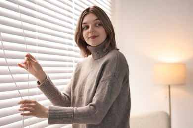 Photo of Young woman adjusting window blinds at home
