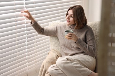 Photo of Woman with cup of drink sitting on armchair near window blinds indoors