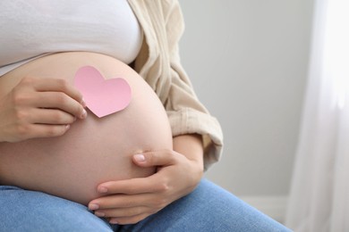 Photo of Pregnant woman with pink paper heart at home, closeup. Space for text