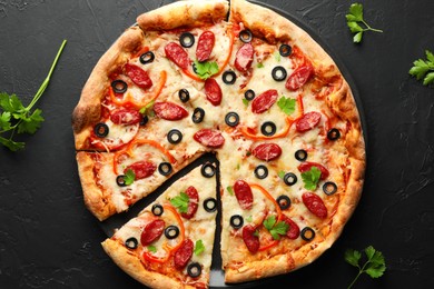 Photo of Tasty pizza with dry smoked sausages, olives, pepper and parsley on black table, top view