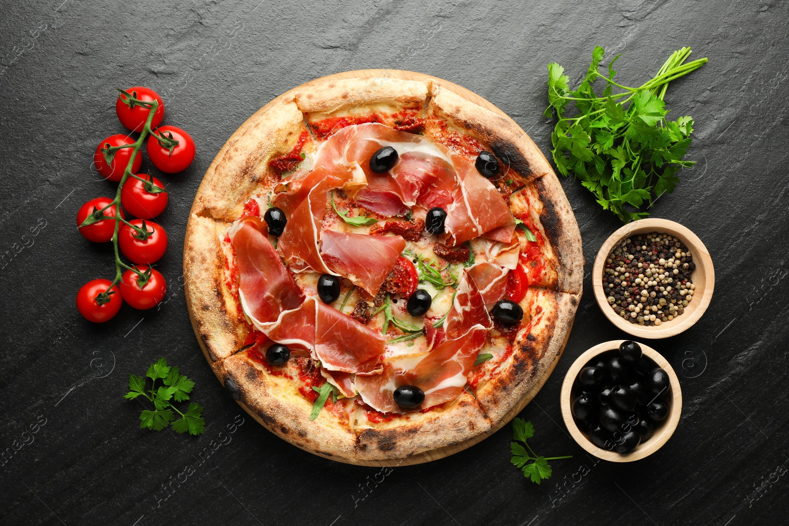 Photo of Tasty pizza with cured ham, olives, tomatoes, parsley and spices on black table, top view