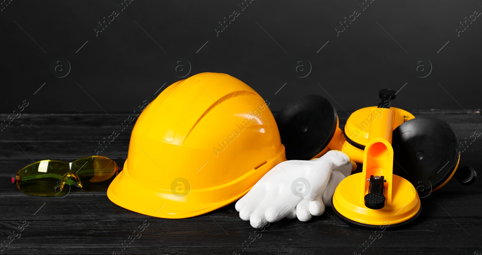 Photo of Hard hat, suction lifters, protective gloves and goggles on black wooden surface against gray background