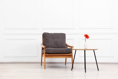 Photo of Comfortable armchair and coffee table with gerbera flower near white wall. Interior design