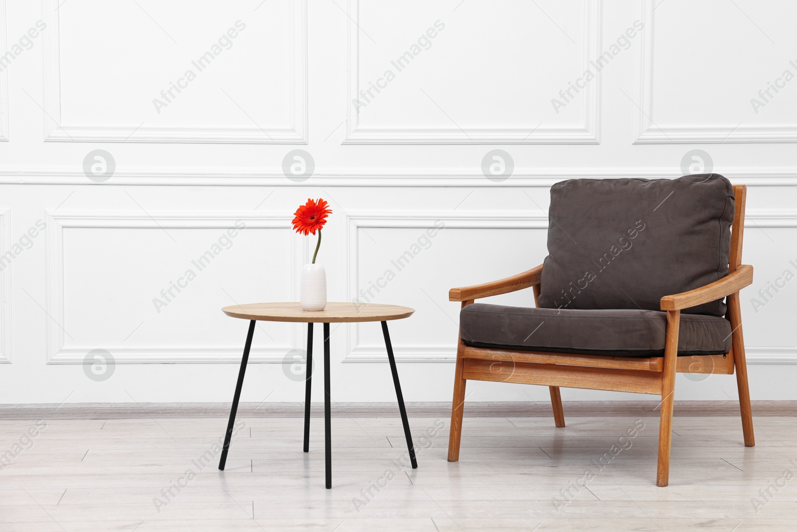 Photo of Comfortable armchair and coffee table with gerbera flower near white wall. Interior design