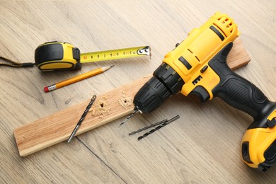 Cordless electric drill, pencil, bits, measuring tape and plank on wooden table, flat lay