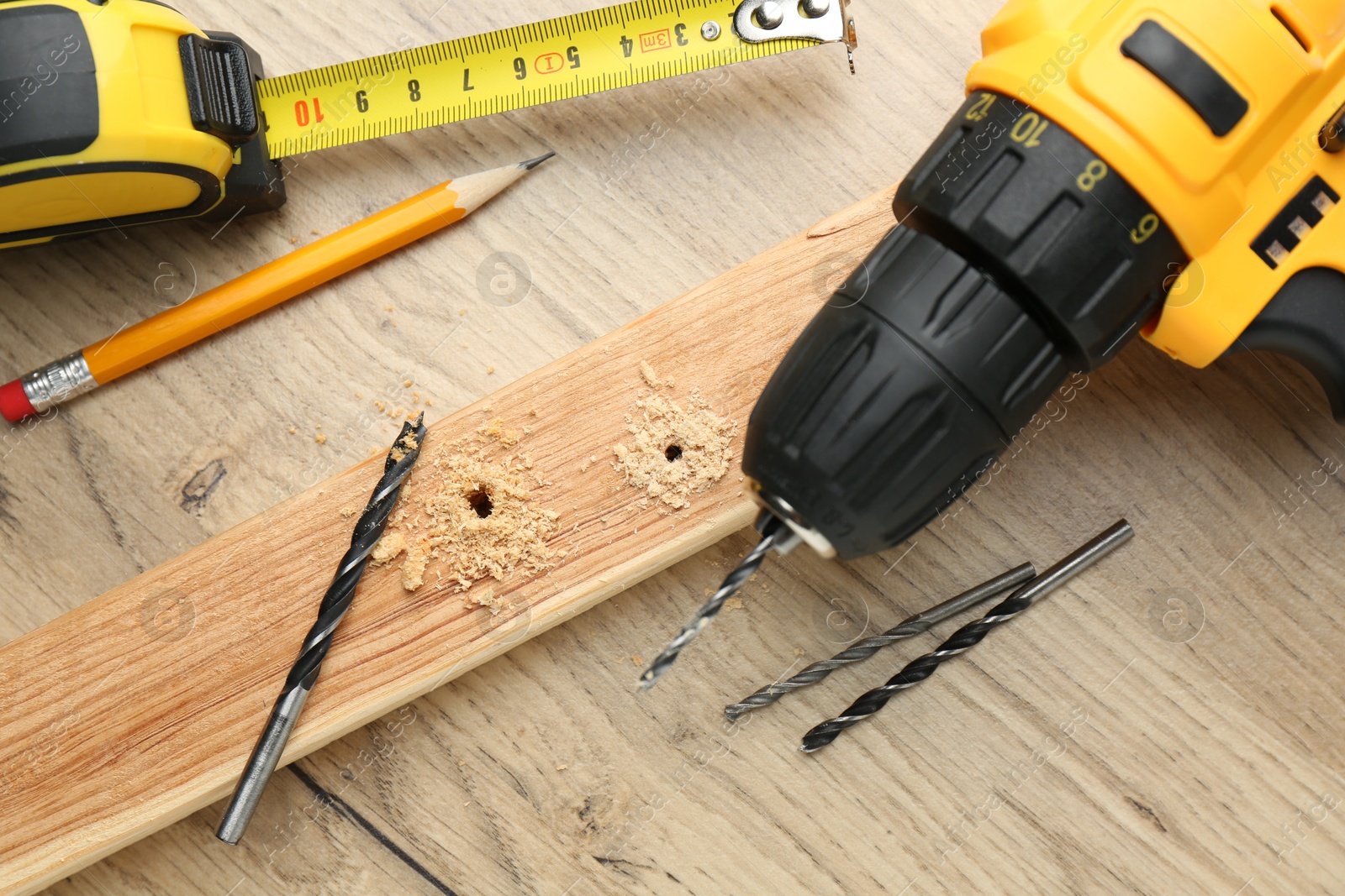 Photo of Cordless electric drill, measuring tape, pencil, bits and holed plank on wooden table