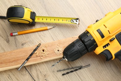 Cordless electric drill, pencil, bits, measuring tape and plank on wooden table, flat lay