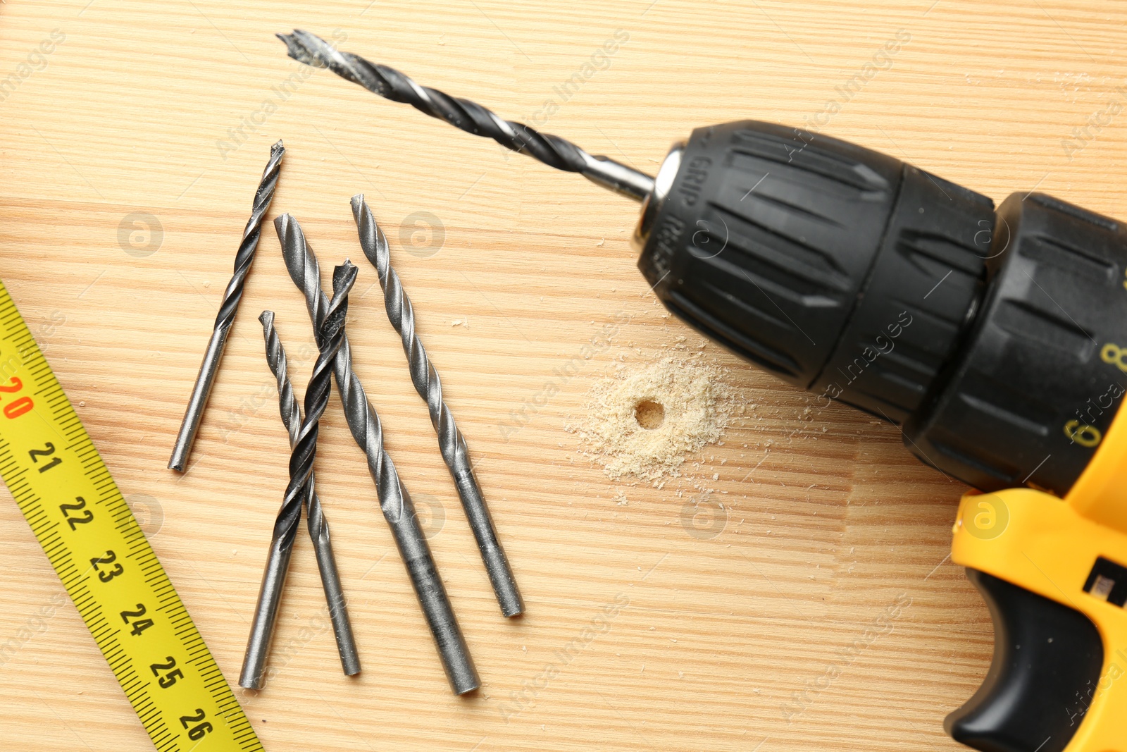 Photo of Cordless electric drill, bits and measuring tape on holed wooden surface, flat lay