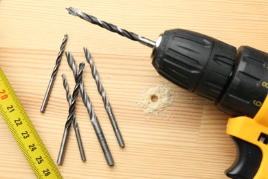 Photo of Cordless electric drill, bits and measuring tape on holed wooden surface, flat lay