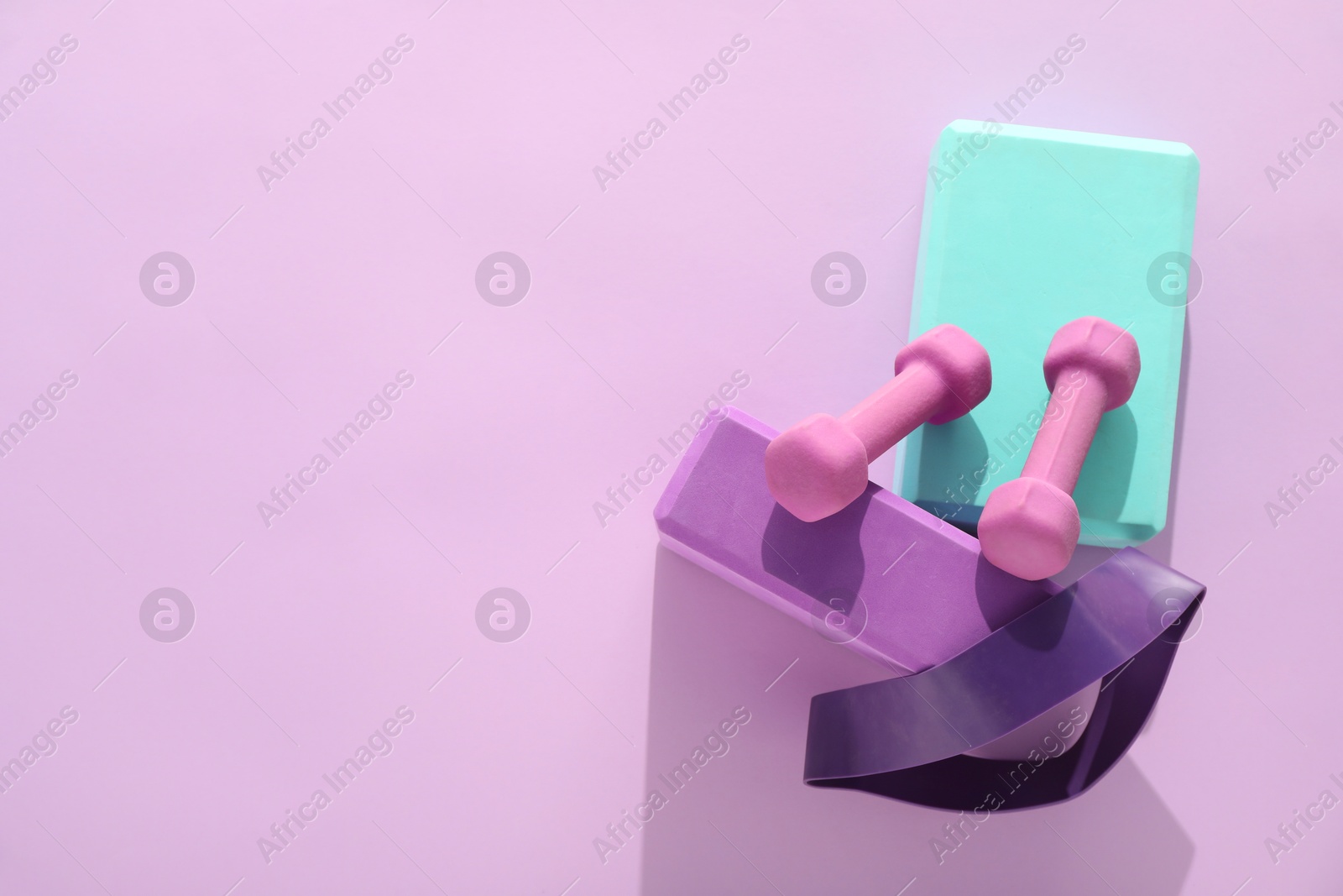 Photo of Two dumbbells, yoga blocks and fitness elastic band on violet background, flat lay. Space for text