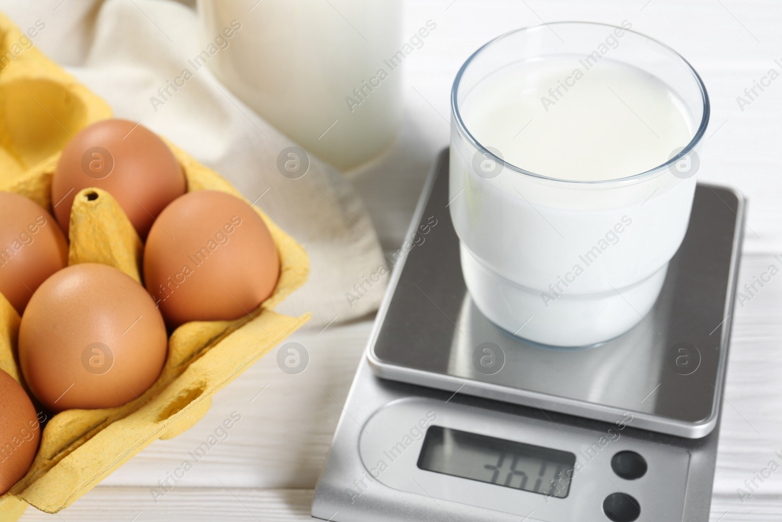 Photo of Kitchen scale with glass of milk near eggs on white wooden table, closeup