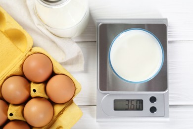 Photo of Kitchen scale with glass of milk near eggs on white wooden table, flat lay