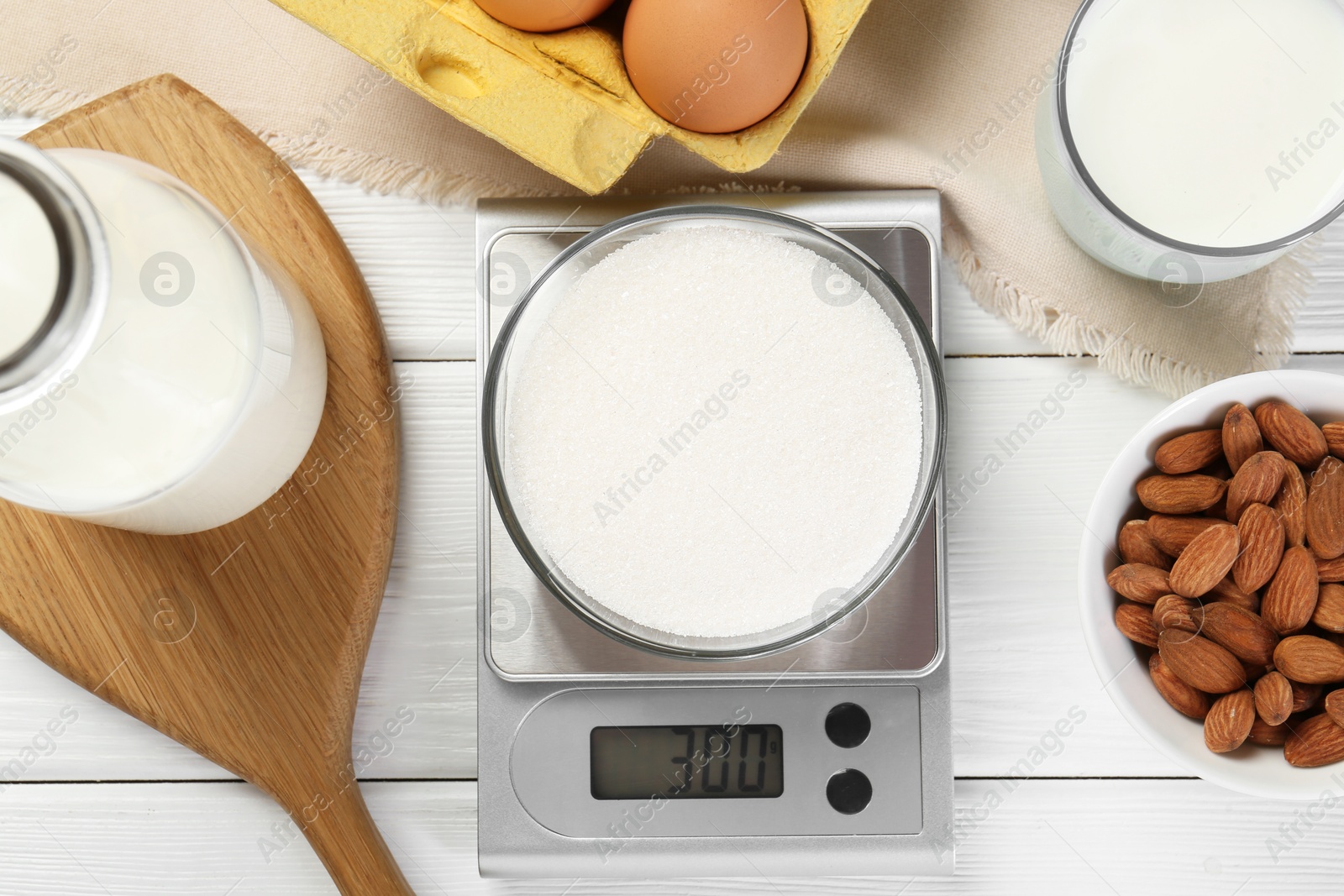 Photo of Kitchen scale with bowl of sugar among milk, almond and eggs on white wooden table, flat lay