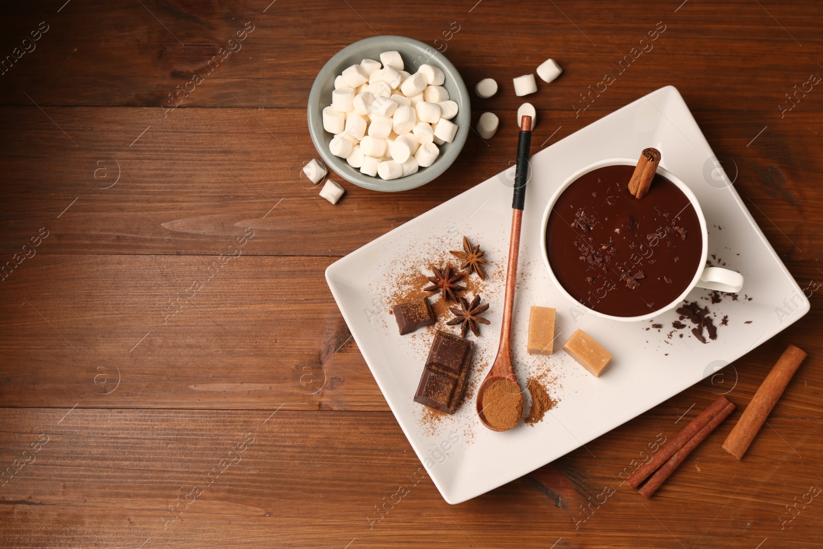 Photo of Tasty melted chocolate in cup, marshmallows, candies and spices on wooden table, flat lay. Space for text